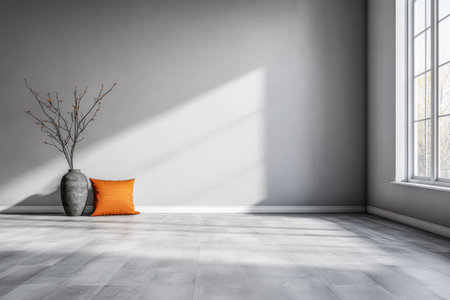A minimalistic interior space features a gray vase with decorative branches and a bright orange pillow on the floor, illuminated by soft sunlight streaming through large windows.の素材