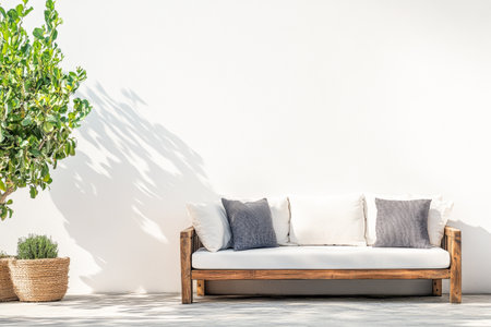 A stylish wooden couch with gray and white pillows is positioned against a clean white wall, accompanied by vibrant greenery basking in the sunlight.の素材