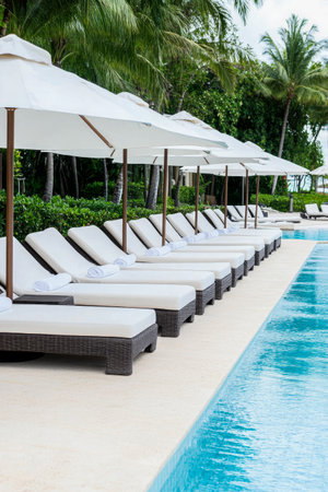 The elegant lounge chairs, arranged side by side, invite guests to unwind by the sparkling pool, shaded by large umbrellas against a tropical backdrop.の素材