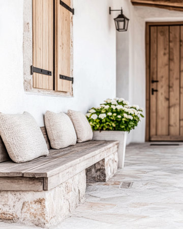 A rustic wooden bench adorned with soft pillows invites relaxation outside a charming entrance, complemented by vibrant white flowers in a pot nearby.の素材