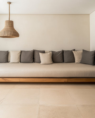 A sleek lounge area showcases a low-profile sofa adorned with beige and gray pillows, complemented by a natural woven pendant light hanging above, creating a serene atmosphere.の素材
