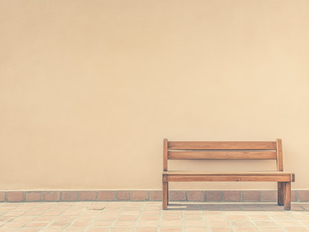 A simple wooden bench sits empty on a tiled floor, positioned in front of a plain beige wall, creating a peaceful and minimalistic outdoor setting under bright daylight.の素材