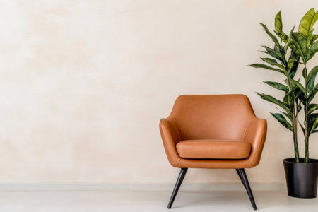 A modern brown leather chair is positioned beside a lush green potted plant, enhancing the minimalist aesthetic of the light-colored wall in a cozy living area.の素材