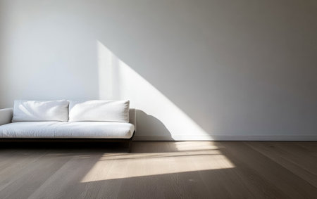 A sleek white sofa sits against a minimalist wall in a modern living room. Bright afternoon sunlight streams through, creating a beautiful shadow on the wooden floor.の素材