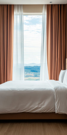 A well-lit hotel room features a comfortable bed beside large windows with flowing curtains, revealing a stunning landscape of hills under a cloudy sky.の素材