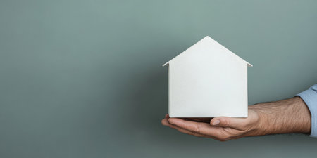 A hand gently holds a small white house model, showcasing its simple design against a soft gray wall. The setting suggests a contemporary atmosphere, focusing on home and comfort.の素材
