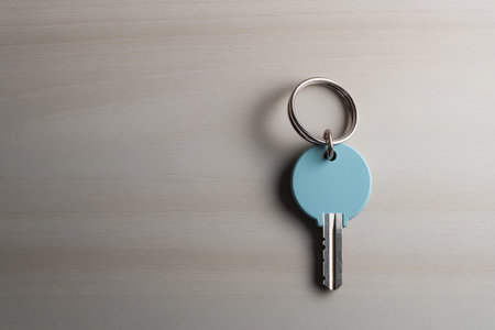 A blue key attached to a silver ring lies on a light wooden surface, illuminated by gentle natural light, creating a calm and minimalistic atmosphere.の素材