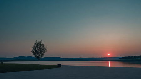 The sun sets gently on the horizon, casting a warm glow over tranquil waters. A solitary tree stands nearby, framed against distant hills, creating a peaceful atmosphere.の素材