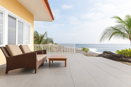A comfortable seating area features a sofa and table on a terrace by the sea, surrounded by palm trees and pebbly shore. The calm ocean waves reflect the blue sky, creating a tranquil atmosphere.の素材