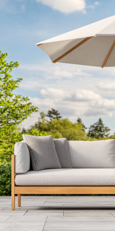 A light gray sofa with cushions is placed outdoors under a large umbrella, surrounded by lush green trees. The bright sky adds a peaceful ambiance to the relaxing space.の素材