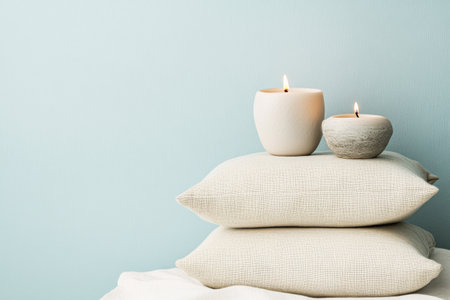 Soft, neutral pillows are stacked neatly as two lit candles add warmth and tranquility to the serene setting against a light blue wall.の素材