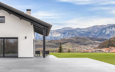 A contemporary home features large windows and a spacious patio overlooking breathtaking mountains. The peaceful landscape includes vibrant greenery and distant village scenery.の素材