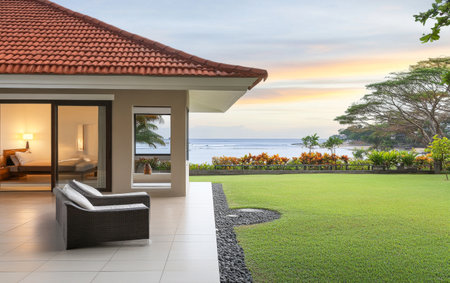 A modern beachfront villa offers a comfortable outdoor seating area overlooking lush greenery and a serene ocean. The beautiful sunset casts a warm glow across the landscape.の素材
