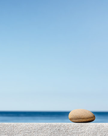 A solitary, smooth pebble is positioned on a stone ledge, with a vast ocean stretching into the horizon beneath a clear blue sky. The peaceful setting invites contemplation and tranquility.の素材
