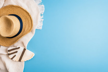A straw hat rests beside a white towel, accompanied by a seashell, all set against a bright blue backdrop. The arrangement evokes a relaxing day at the beach, perfect for summer vibes.の素材