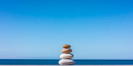 Smooth, round stones are carefully stacked in a balanced formation on a ledge, with a serene blue ocean and clear sky in the background, creating a peaceful, calming atmosphere.の素材