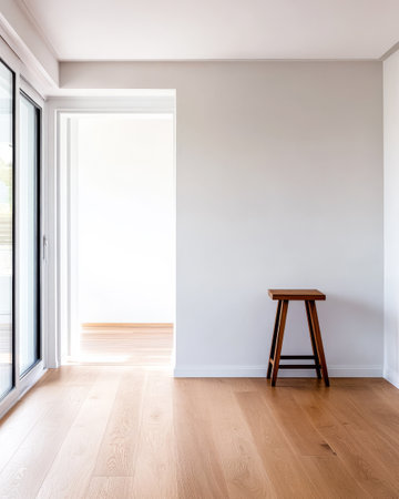 A bright and airy room features a simple wooden stool against a light gray wall. Natural light floods in through large windows, highlighting the wooden floor and creating a serene atmosphere.の素材