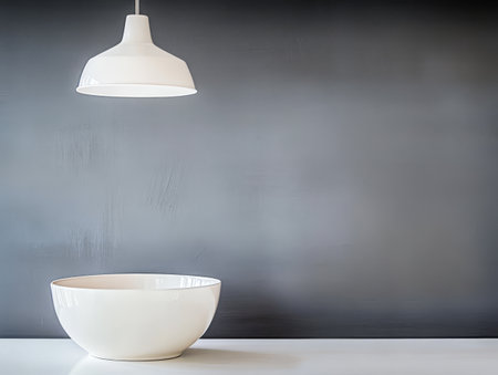 A modern kitchen showcases a sleek white bowl placed on a countertop beneath a stylish pendant light. The gray wall provides a simple and elegant backdrop to the arrangement.の素材
