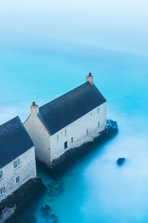 Two stone houses sit on a rocky outcrop surrounded by crystal clear turquoise water. The early morning light casts a serene ambiance highlighting the rugged beauty of the coastline.の素材