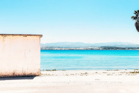 Bright daylight showcases a tranquil beach setting with a white wall on the left turquoise water gently lapping at the shore and distant hills visible on the horizon.の素材