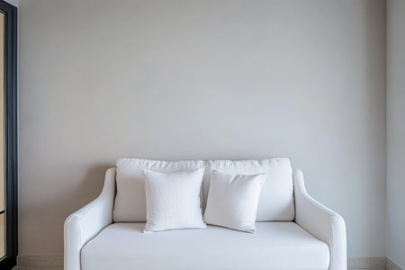 A simple white couch adorned with two decorative pillows rests against a light gray wall creating a serene and minimalist living space.の素材