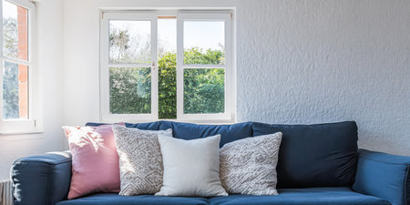 A comfortable blue couch with various decorative pillows is positioned against a white wall. Natural light streams through a large window illuminating the space and greenery outside.の素材