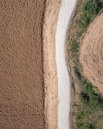 A gravel path winds through farmland bordered by plowed earth on one side and unplanted land on the other. The view captures the texture of the soil and vegetation.の素材