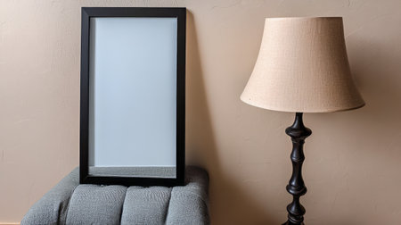 A blank black frame stands upright next to a beige lampshade on a decorative lamp. The setting features a soft gray surface adding a cozy touch to the overall aesthetic.の素材