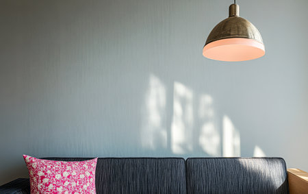 A modern living room features a dark couch with a colorful pillow illuminated softly by a pendant light. Afternoon sunlight casts gentle shadows on the wall.の素材