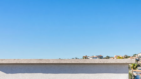 A stunning clear blue sky stretches above a coastal town showcasing a peaceful rooftop view. The white wall in the foreground contrasts with the vibrant landscape.の素材