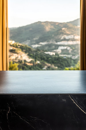 A sleek dark counter stands in a bright room with large windows revealing a stunning mountainous landscape. Green hills and distant buildings create a picturesque setting.の素材