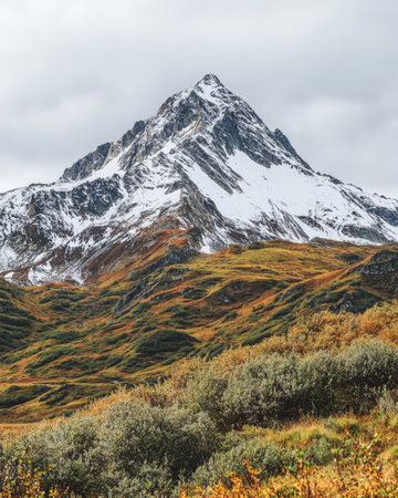 Majestic mountain rises sharply against a backdrop of deep green and vibrant autumn colors with snow-capped summit contrasting the rich landscape under a cloudy sky.の素材