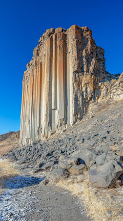 Towering basalt columns rise sharply from the rocky terrain showcasing their unique geometric formations against a bright blue sky surrounded by sparse vegetation and rugged ground.の素材