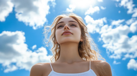A young woman with wavy hair gazes upwards basking in the sunlight on a clear day. Fluffy clouds drift across a vivid blue sky enhancing the serene atmosphere.の素材