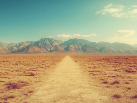 A long dusty path stretches into the distance surrounded by arid land and sparse vegetation. Majestic mountains rise in the background against a serene blue sky.の素材