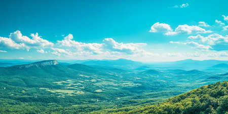 Expansive view features rolling green hills and distant mountains beneath a bright blue sky. Fluffy clouds drift across the horizon emphasizing the natural beauty of the landscape.の素材