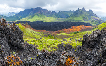 Vibrant green hills surround a vast black lava field showcasing the powerful geological forces at play in this volcanic area. The dramatic mountains loom in the distance under a cloudy sky.の素材