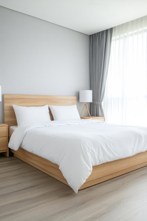 A spacious bedroom showcases a wooden bed with white linens. Soft light filters through sheer curtains illuminating the minimalistic decor and wooden furniture next to the bed.の素材
