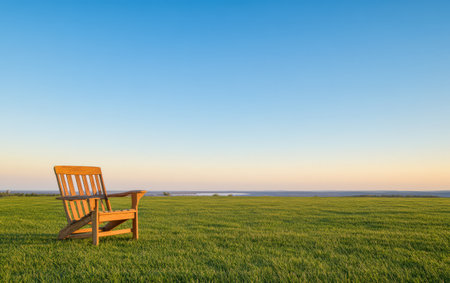A wooden chair stands alone on lush green grass facing a vast open space under a clear blue sky as the sun begins to set in the distance creating a peaceful and inviting atmosphere.の素材