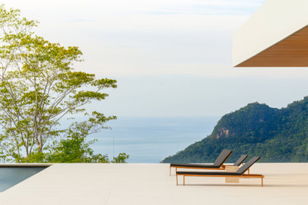 Two lounge chairs sit on a spacious terrace overlooking a peaceful ocean and lush green mountains. The serene setting is perfect for unwinding in a modern architectural design.の素材