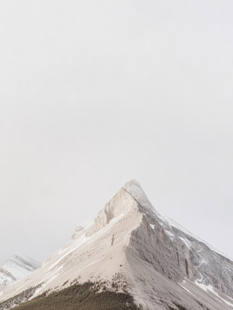 A stark mountain peak rises sharply against a pale sky partially shrouded in clouds. Lush green trees lie at the base creating a serene contrast to the rugged terrain.の素材