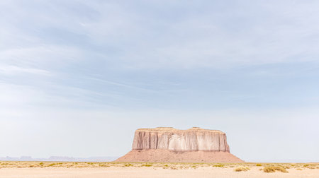 A striking rock formation stands prominently on the arid landscape of Monument Valley. The clear sky enhances the beauty of this iconic natural landmark surrounded by bare land.の素材