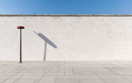 A simple urban landscape shows a solitary post casting a long shadow against a clean white wall under a clear blue sky. The setting highlights minimalism in everyday architecture.の素材