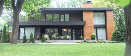 A contemporary house features sleek lines and a unique wooden facade set in a vibrant garden with manicured lawns and various plants. Large windows illuminate the interior.の素材