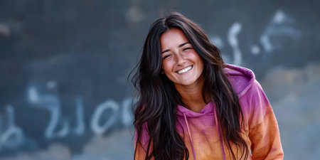 A young woman with long dark hair wears a vibrant tie-dye hoodie and smiles warmly. She stands outdoors radiating joy against a textured graffiti-covered wall in a lively urban environment.の素材