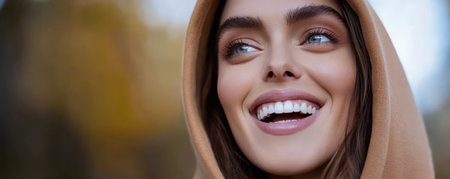 A young woman wearing a brown hoodie smiles brightly while outdoors. The autumn sun casts a warm glow around her highlighting her joyful expression amidst soft blurred colors of nature.の素材