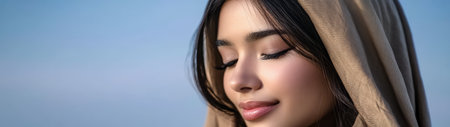 A young woman with long dark hair is wearing a light brown headscarf. She appears peaceful with her eyes closed and a gentle smile basking in warm natural light outdoors.の素材