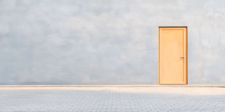 A bright orange door stands prominently on a plain gray wall creating a striking contrast. The surrounding area features a clean paved surface illuminated by soft daylight.の素材