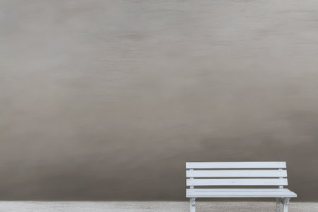 A white wooden bench sits alone in front of a smooth gray wall, creating a serene atmosphere. The simplicity of the setting invites rest and contemplation in this open area.の素材
