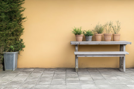 A rustic wooden bench displays several potted plants atop it, set against a bright yellow wall. The area is serene with a touch of greenery from nearby foliage.の素材